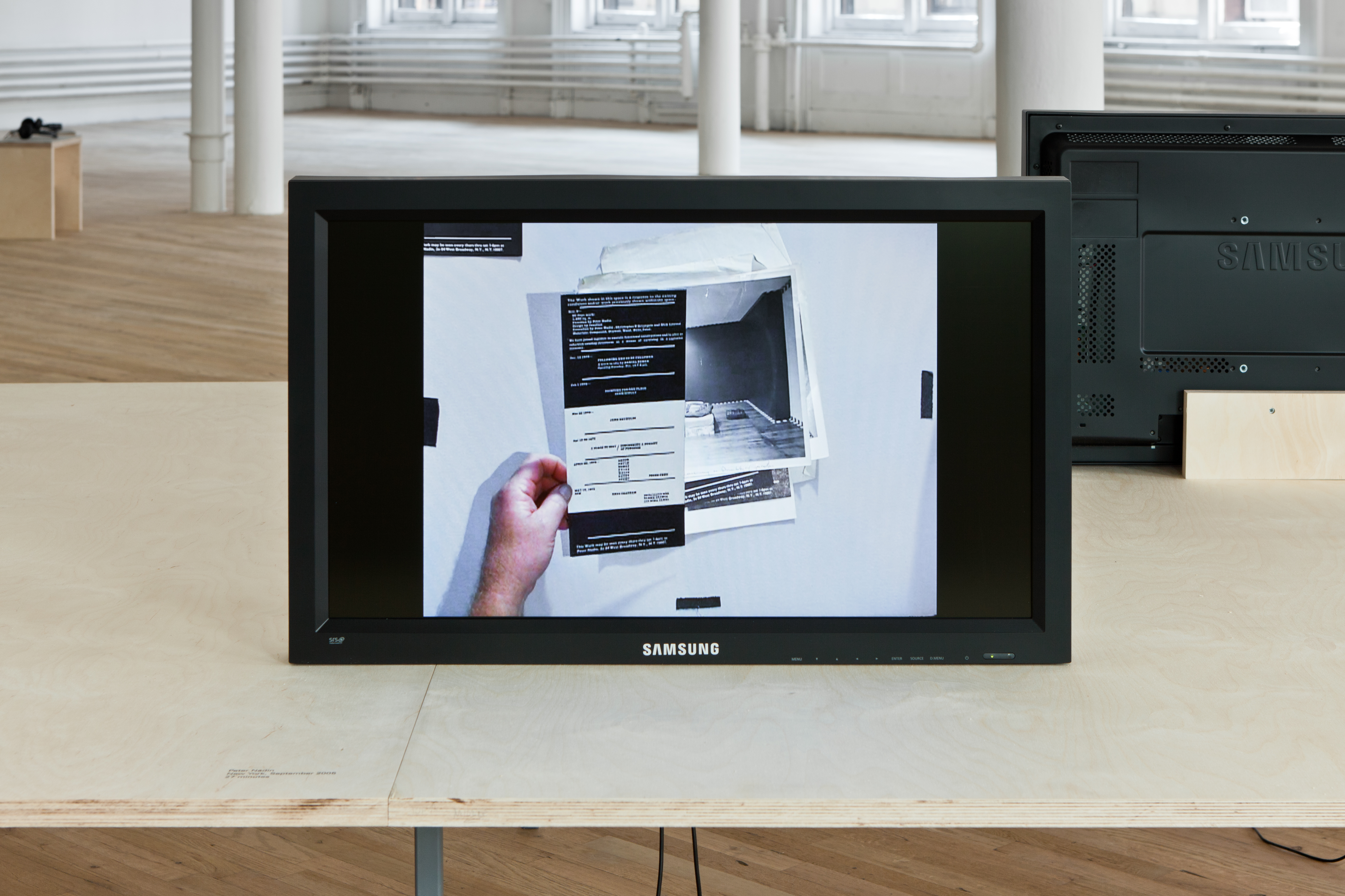 A black monitor sits on a wooden table. The image onscreen shows a hand holding a black and white paper with text. A stack of photographs lay underneath the paper.