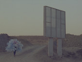 A color film still depicting a person who walks up a dirt path carrying a myriad of empty plastic bottles. To the person's right is a billboard facing away from the camera.