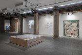 Photograph of a basement gallery space with various multimedia works displayed. In the center is a large wooden platform placed on the floor. Along the back wall is a projected video, a grid of works on paper, and a woven hanging piece.