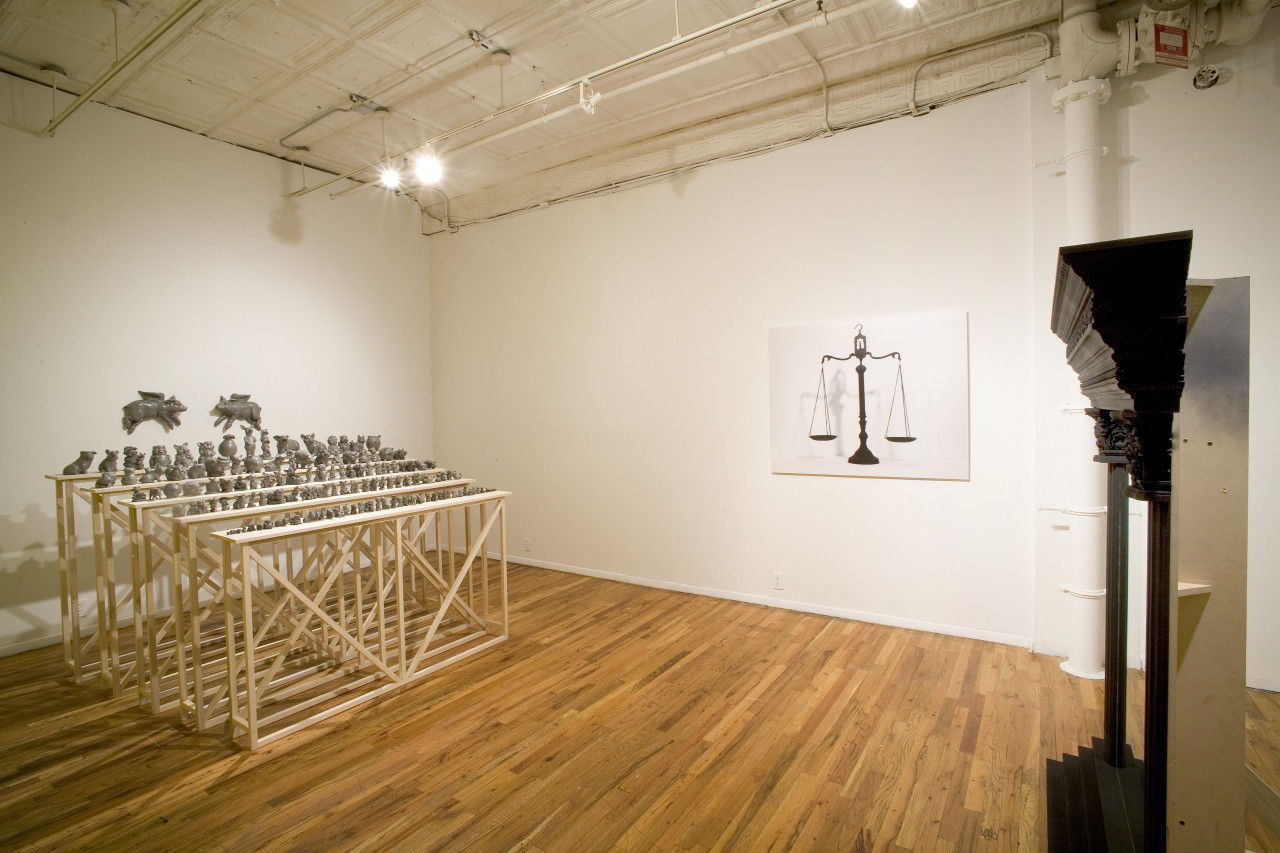 Five rows of bleachers built from wood, with dozens and dozens of assorted sculptures of gray, painted pigs arranged in decreasing size from the top bleacher to the bottom. Two flying pigs are hung on the wall directly behind the bleachers. Several feet away, across from the pigs, is a side view of a frame made of columns. On the wall between the pigs and the columns is a poster of hanging scales.