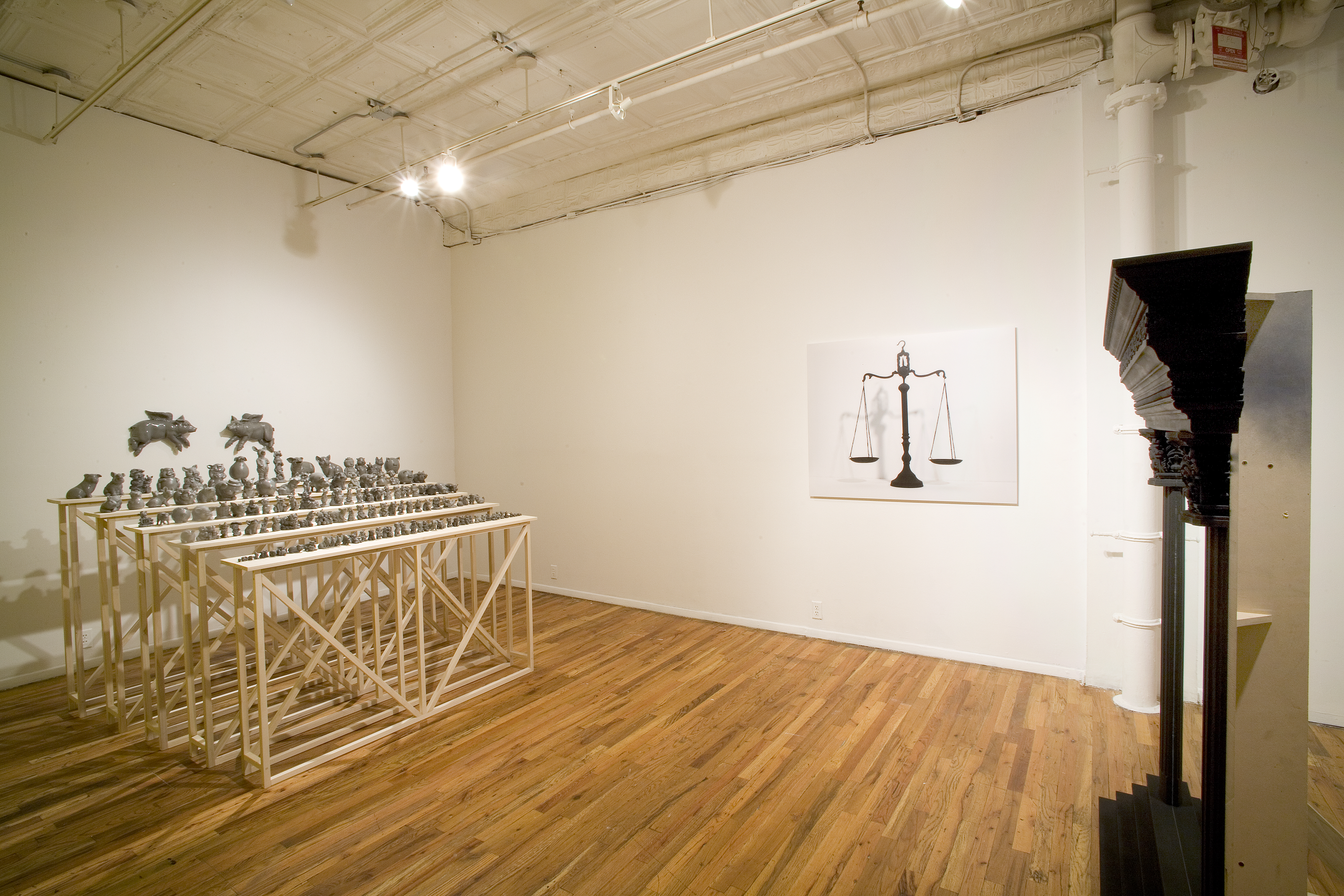 Five rows of bleachers built from wood, with dozens and dozens of assorted sculptures of gray, painted pigs arranged in decreasing size from the top bleacher to the bottom. Two flying pigs are hung on the wall directly behind the bleachers. Several feet away, across from the pigs, is a side view of a frame made of columns. On the wall between the pigs and the columns is a poster of hanging scales.