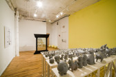 Five rows of bleachers built from wood, with dozens and dozens of assorted sculptures of gray, painted pigs. The pigs are reflected in a mirror opposite them, which is framed by columns. On a wall on the left side is an image of hanging scales. The right wall is painted yellow.