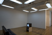 In a gallery space, a large white overhead LED-style sign acts as both a drop ceiling and overhead lighting. It displays two numbers, a 2 and a 3. Below are a black sofa and, facing it, a small television on a wooden pedestal.