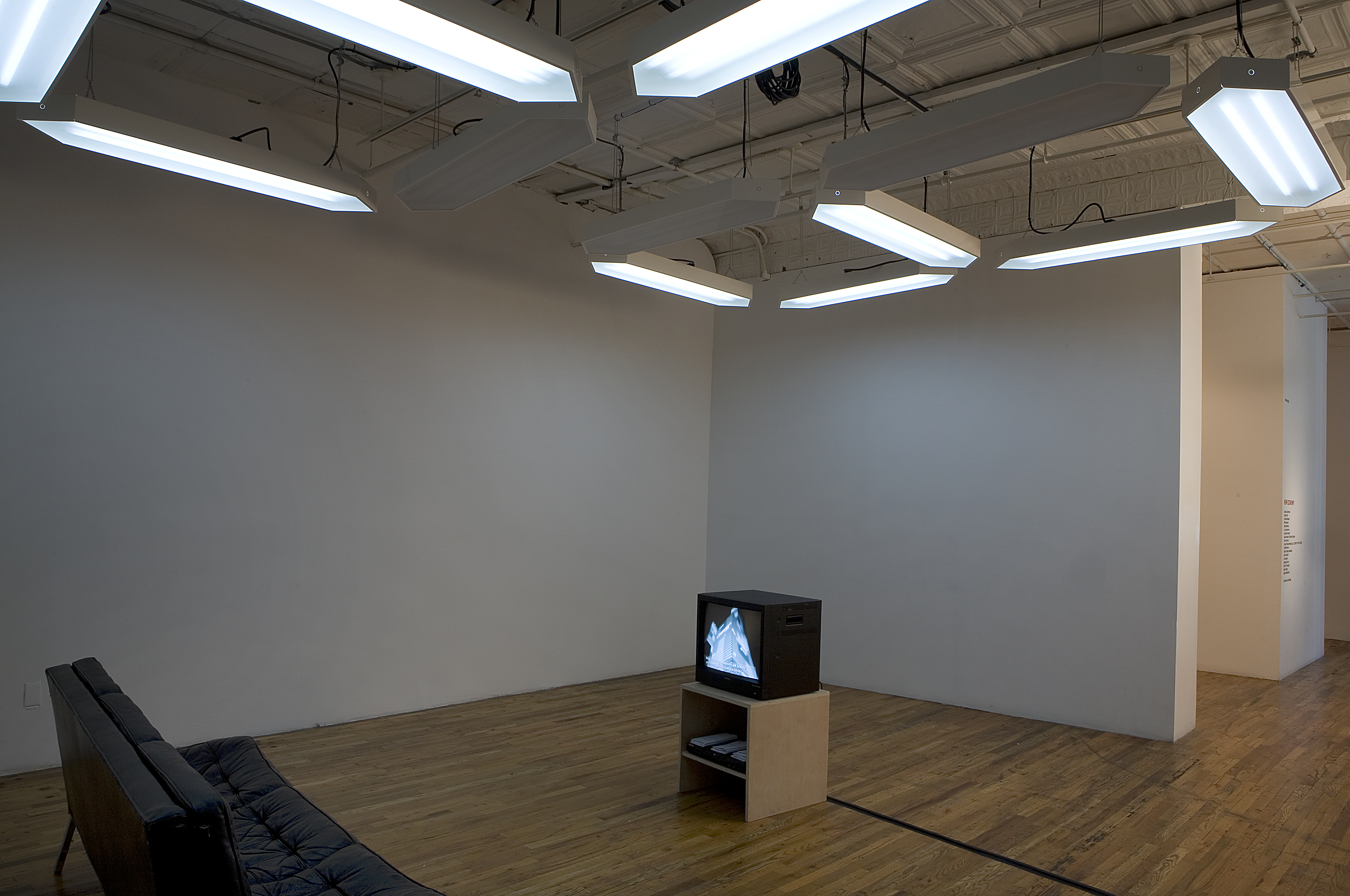 In a gallery space, a large white overhead LED-style sign acts as both a drop ceiling and overhead lighting. It displays two numbers, a 2 and a 3. Below are a black sofa and, facing it, a small television on a wooden pedestal.