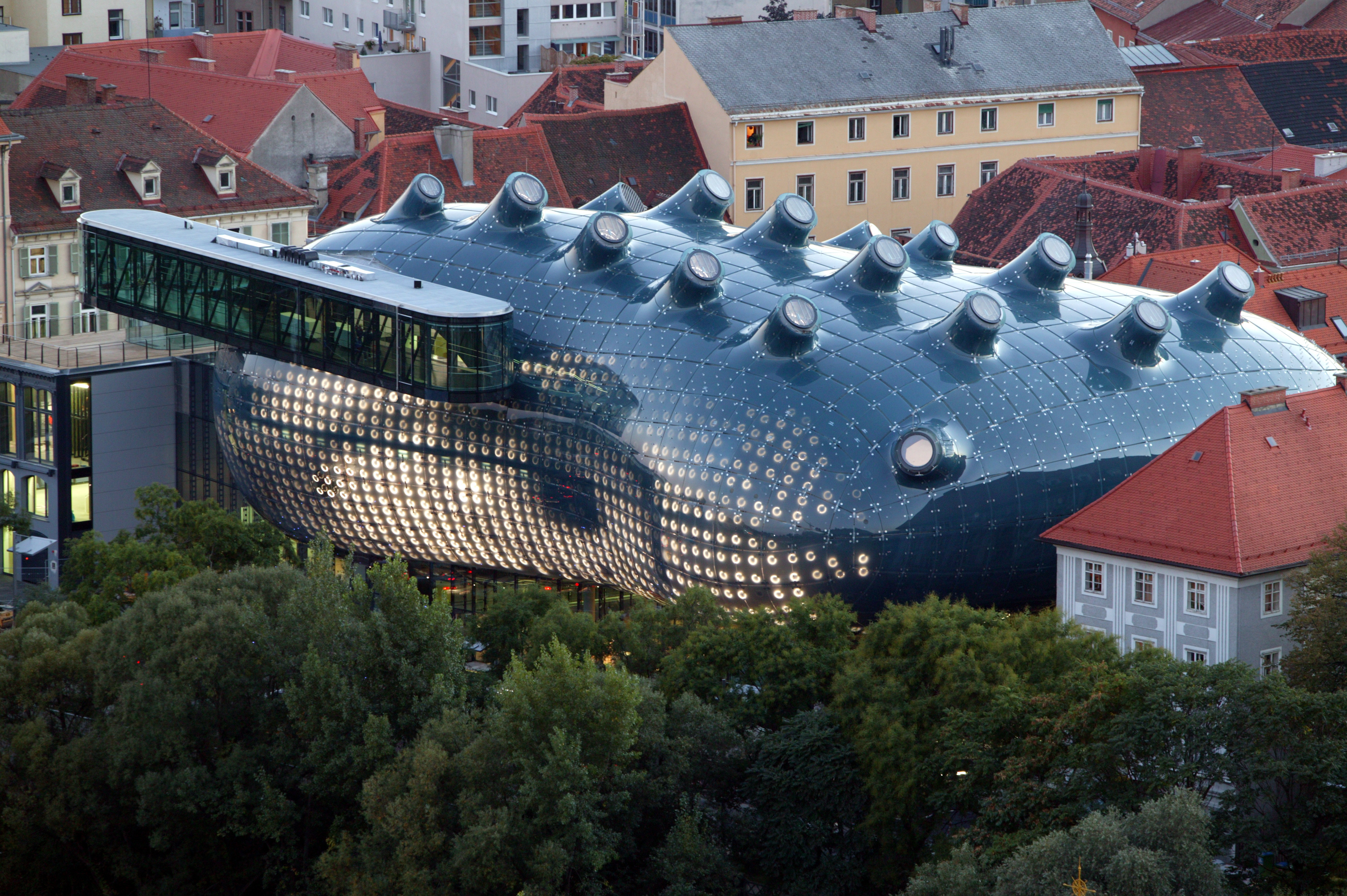 An overhead view of a very modern building with a dark, bulbous, skin-like facade. Protruding from its top are tuberous skylights all facing the same direction, and at left is a long elevated walkway.
