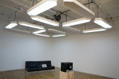 In a gallery space, a large white overhead LED-style sign acts as both a drop ceiling and overhead lighting. It displays two numbers, a 2 and a 5. Below are a black sofa and, facing it, a small television on a wooden pedestal.