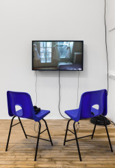 Two blue chairs face a monitor that displays a still image of framed windows in which three curtains have been tied to look like bodies in dresses.