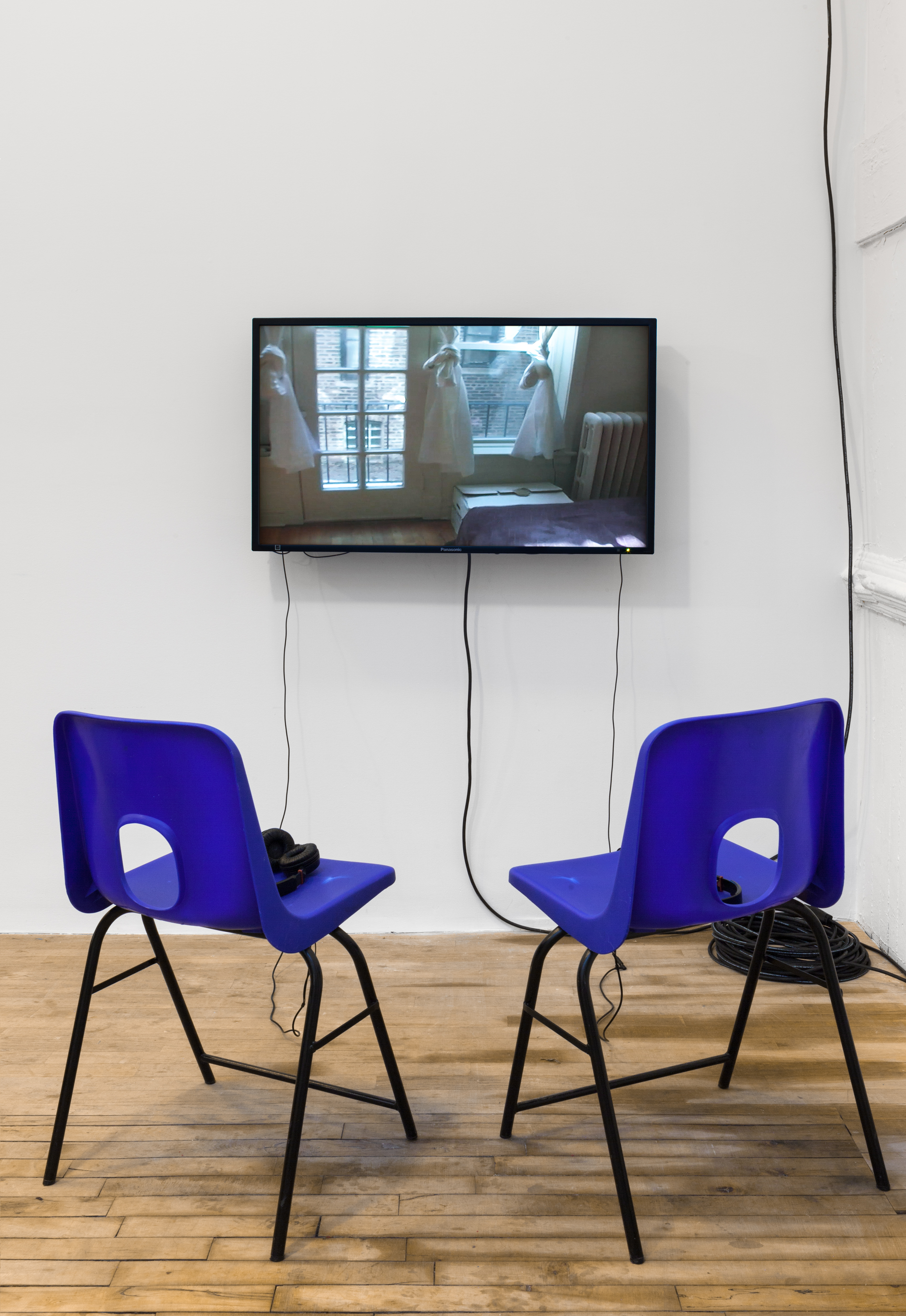 Two blue chairs face a monitor that displays a still image of framed windows in which three curtains have been tied to look like bodies in dresses.