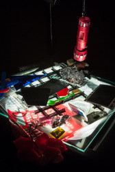 An angled view of the illuminated stage of a overhead projector. A red flashlight descends from the lamp onto a flat pile of objects including photographs, drawings, transparents material, and more.