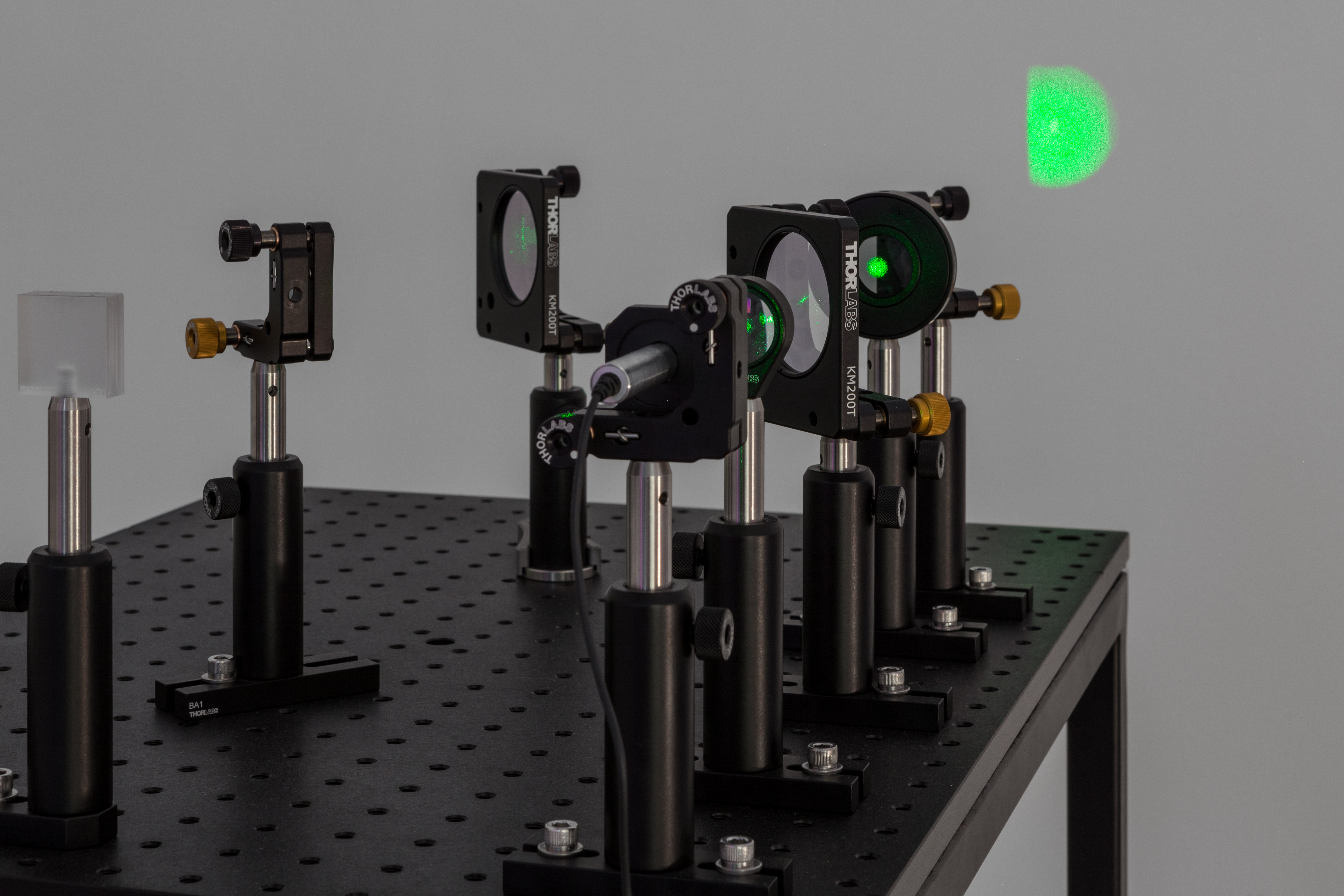 An inferometer in the foreground, bolted onto a black peg-board table, projects a green dot, through multiple lenses, onto a white wall.