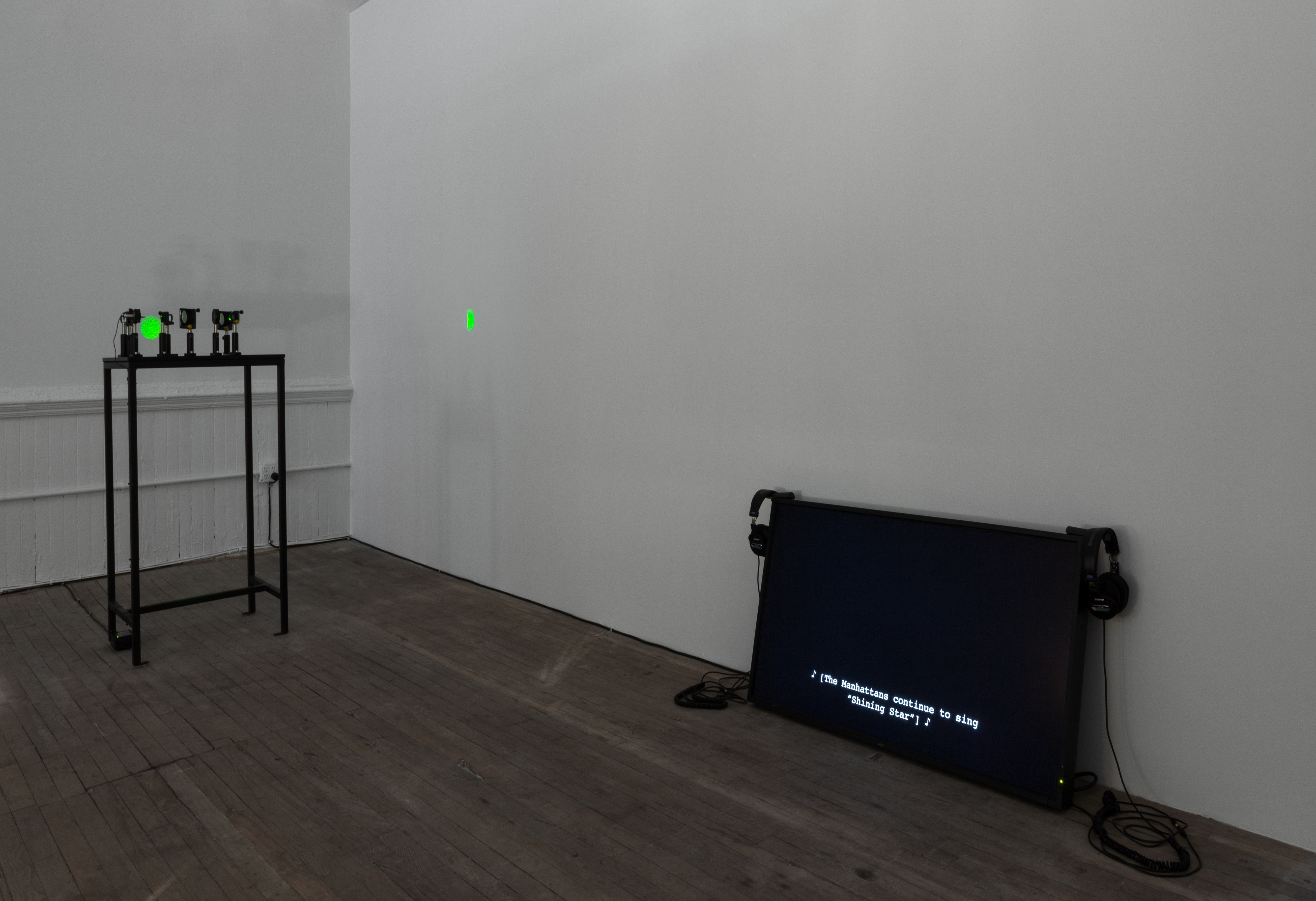 A view of a dimly lit room with a video monitor sitting on the ground of the right wall. The monitor screen is black with white text reading, 