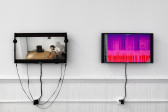 Two monitors are hung on a white wall. On the left, an image of a man crouching in a room with a chair sitting on the ground behind him. On the right, a purple, pink, and red graph is displayed next to a column of indicating numbers.