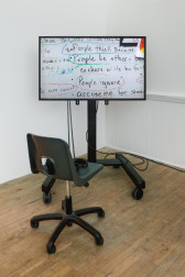 A monitor on a wheeled stand is displayed in the corner of two white walls. The monitor projects an image of a whiteboard with hand written text that reads, 