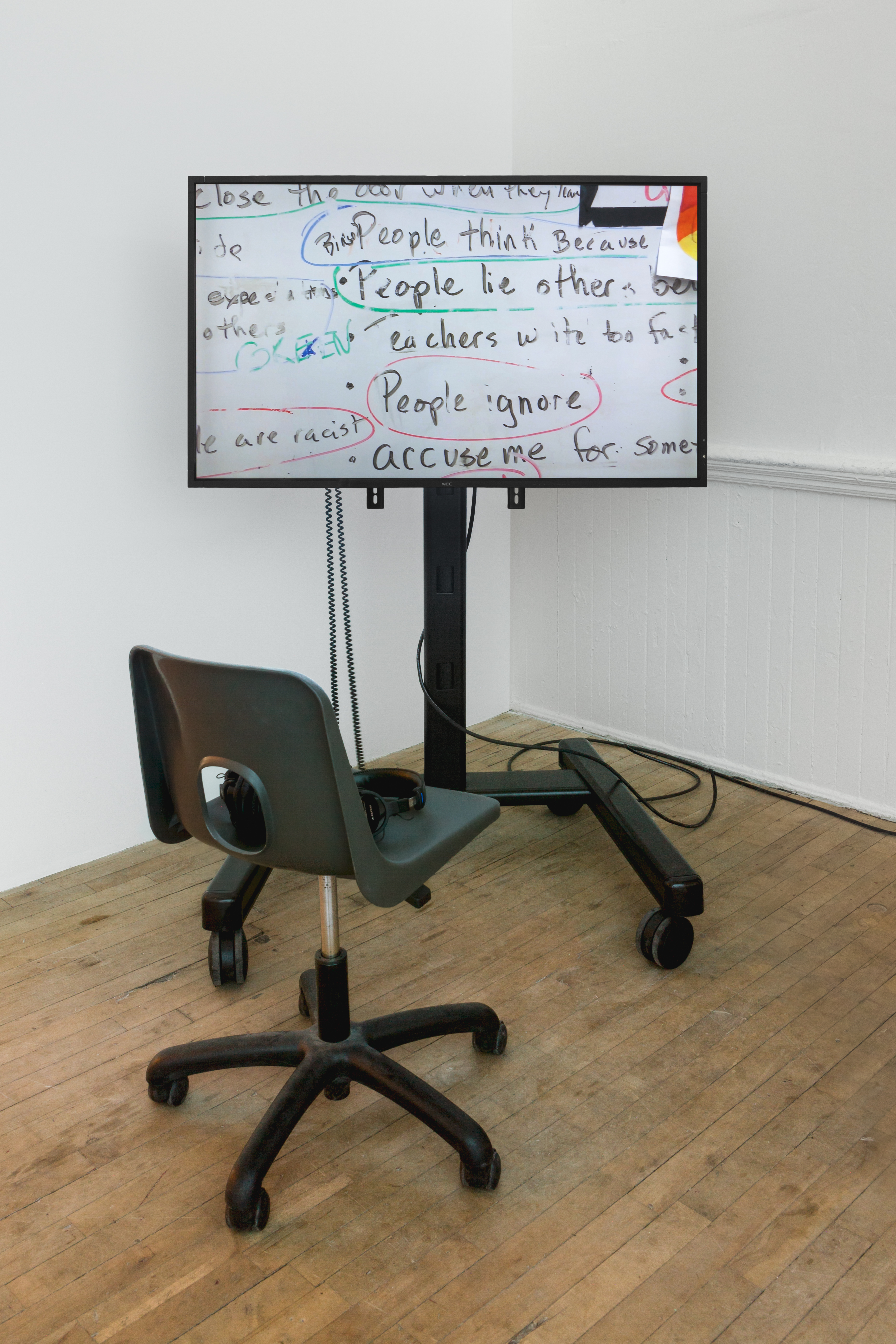 A monitor on a wheeled stand is displayed in the corner of two white walls. The monitor projects an image of a whiteboard with hand written text that reads, 