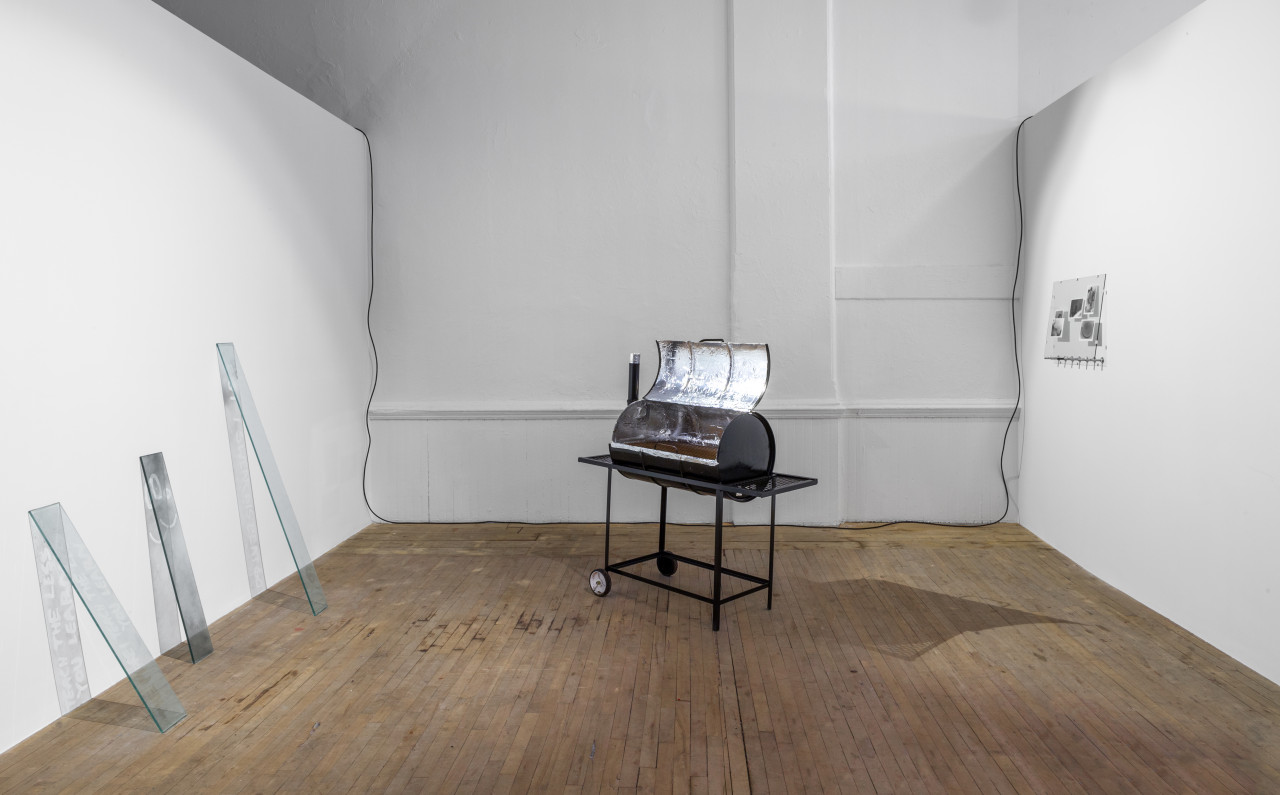 A barrel, converted into a grill, sits horizontally on a wheeled cart. It is opened, with a silver metallic interior, in the center of the room. On the left, three long planks of glass lean onto the wall. To the right, photographs displayed in a pane of glass sit at eye-level on the wall.