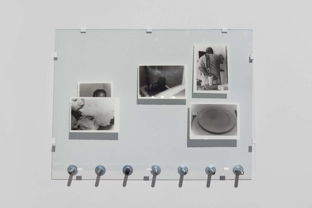 Five photographs arranged in a pane of glass lean on bolts against a wall. The images in the photographs (from left to right) include a man exhaling smoke, a man looking towards the viewer, a man in a bathtub, a man, turned away from the camera, in a hospital gown, and an empty dinner plate.