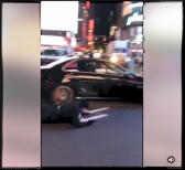 A screencapture from a video taken on a phone in Times Square, depicting a person facedown beside a car, still in motion, in the street.