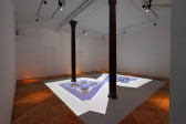 Color image of a video installation in the center of the floor of a gallery. A walkway made of plywood runs along the edges of the room, encircling the projection. The projection depicts a mirrored negative film image. The image features an assortment of abstracted objects arranged on a blue background.