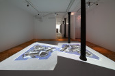 Color image of a video installation in the center of the floor of a gallery space. A walkway made of plywood runs along the edges of the room, around the projection. The projection depicts a negative film image that has been mirrored along the center. Objects are arranged on a blue background to create vaguely recognizable images.