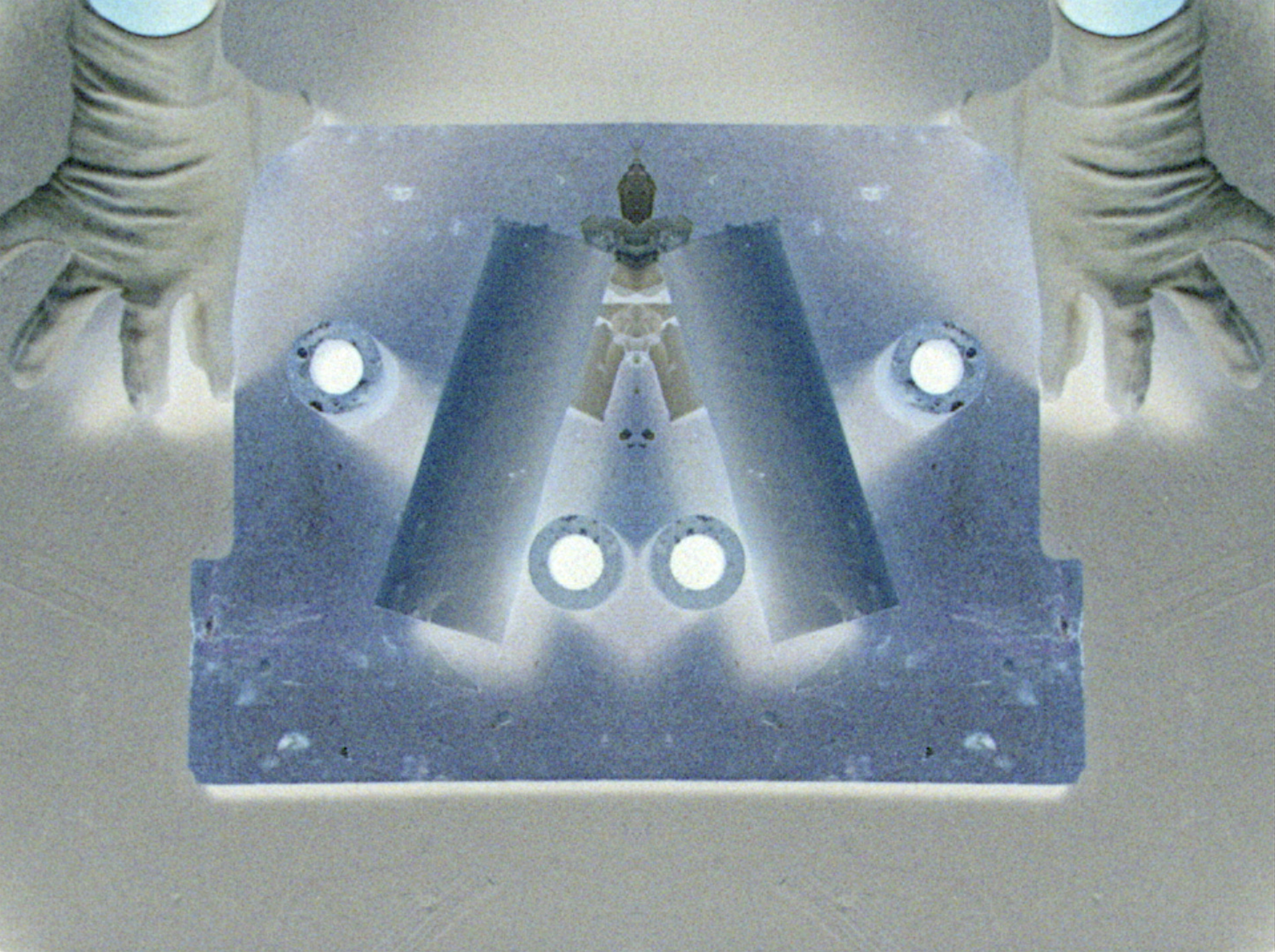 Negative film image of two hands holding down either side of a blue board. Objects are placed to create vaguely recognizable patterns and figures.