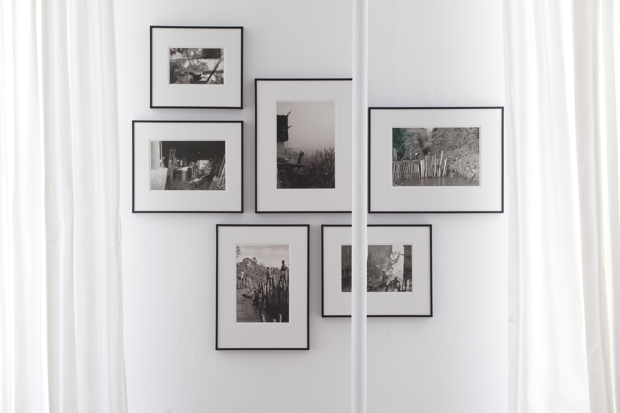 A center-parted white chiffon curtain revealing a set of six framed images mounted on the wall. A white bar runs slightly right of center of the image.