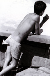 A black and white, vintage photograph of a male figure leaning casually against a low wall by a river. The figure's long back faces the viewer, and we cannot see his face. He wears a short piece of cloth at his waist, while his upper body is bare.