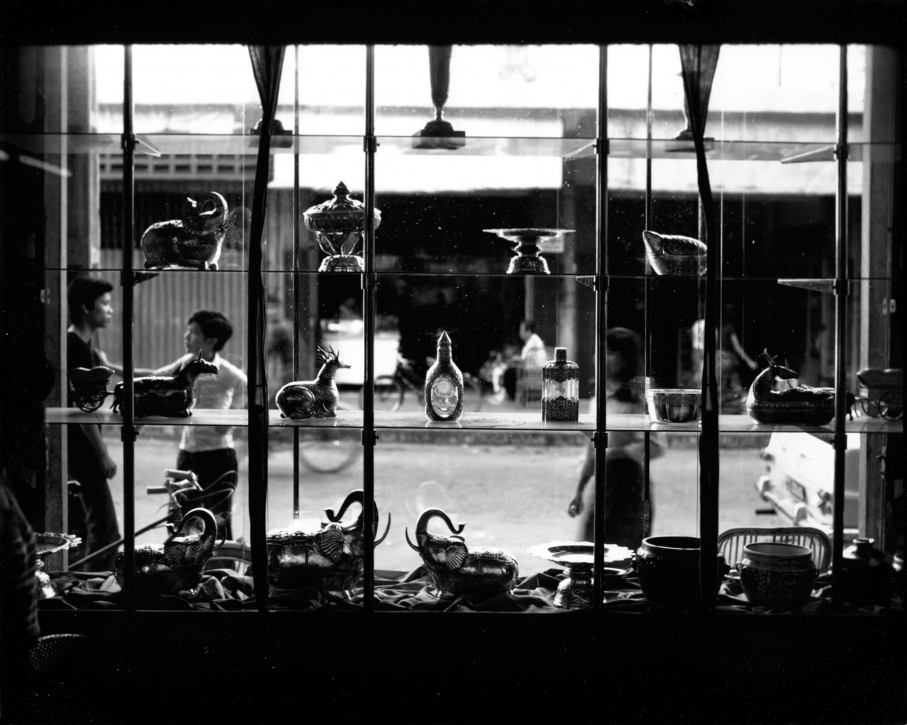 A vintage, black and white photograph of the interior of a shop window, which looks out upon a city street. Two men stand to the left side of the window, casually leaning against a pole and talking. Various metal vases, cutlery, and ornaments are displayed on the shelves within the window.
