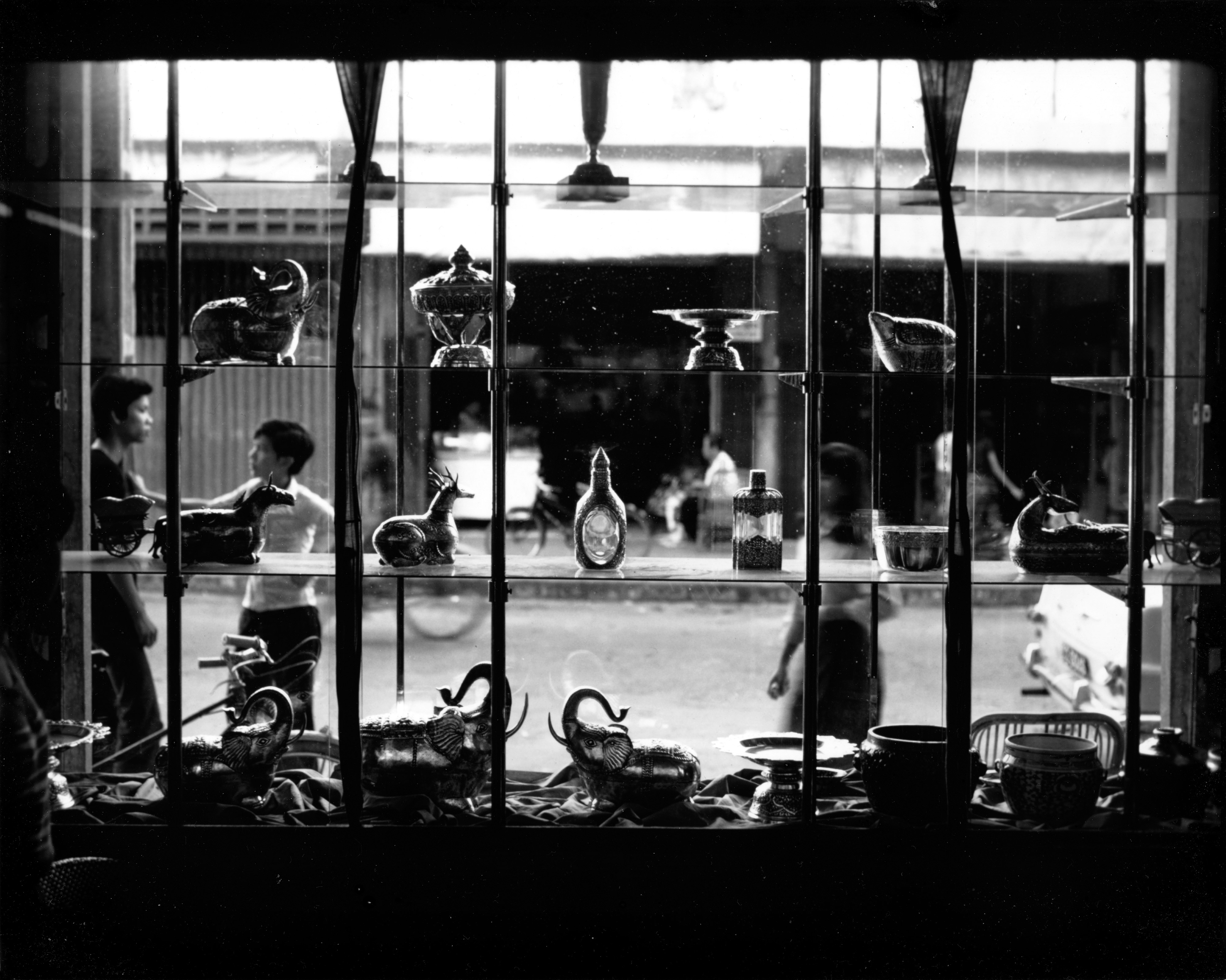 A vintage, black and white photograph of the interior of a shop window, which looks out upon a city street. Two men stand to the left side of the window, casually leaning against a pole and talking. Various metal vases, cutlery, and ornaments are displayed on the shelves within the window.