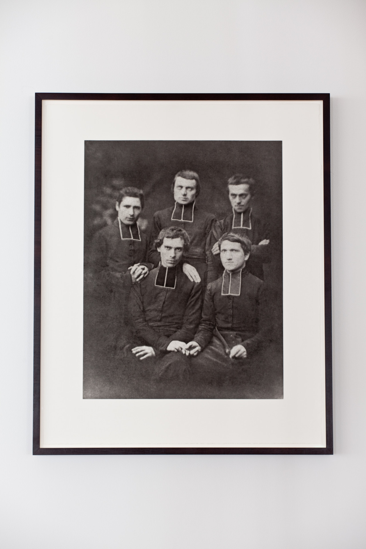 A vintage, framed black and white photograph of five priests dressed in all black stiffly pose before the camera for a portrait. The two men sitting closest to the viewer hold hands.