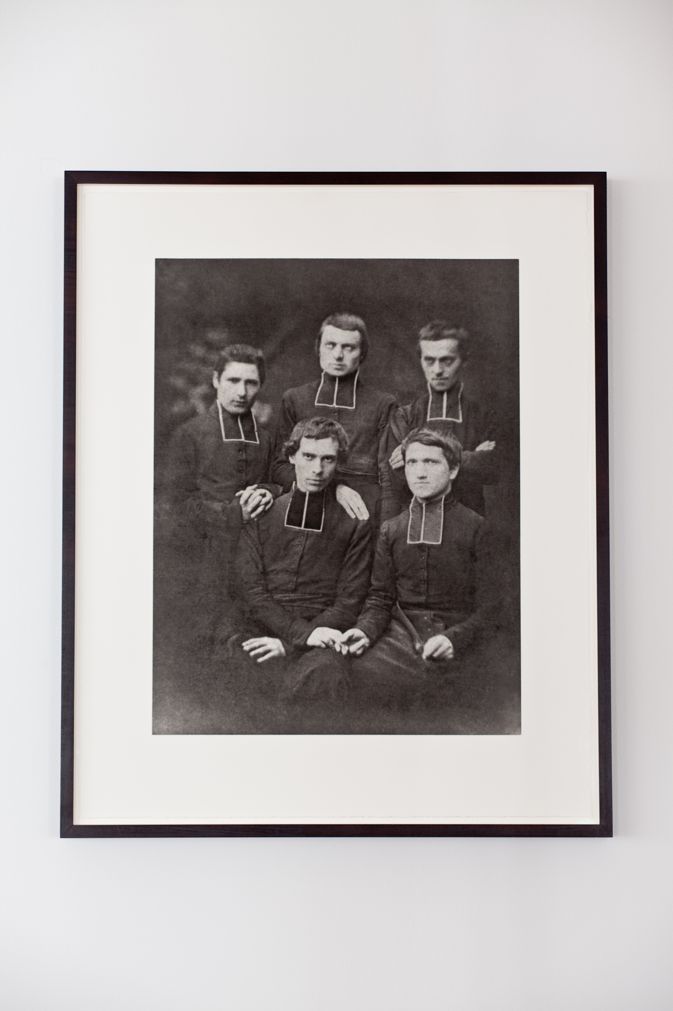 A vintage, framed black and white photograph of five priests dressed in all black stiffly pose before the camera for a portrait. The two men sitting closest to the viewer hold hands.