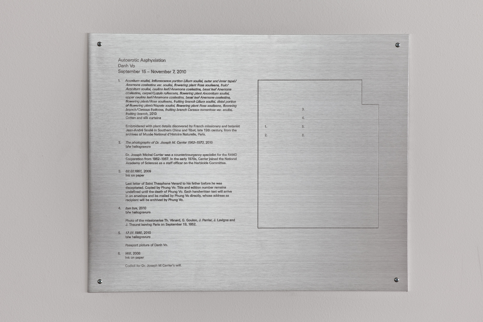 Close-up of a rectangular, silver metal plaque hanging on a white gallery wall. The plaque is inscribed with a diagram of an exhibition space. Numbers note the location of the artworks, while a key next to the diagram notes the titles and other tombstone information of the works.