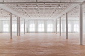 A large exhibition space with light wood flooring, wide windows, and white columns fills with gentle daylight. The far windows and walls the space are covered in white, semi-translucent curtains, which gently glow.