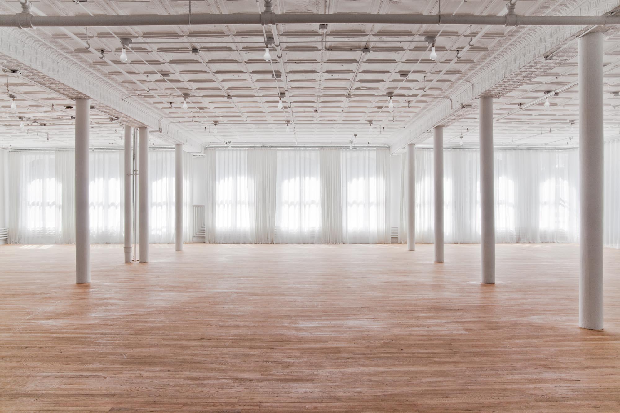 A large exhibition space with light wood flooring, wide windows, and white columns fills with gentle daylight. The far windows and walls the space are covered in white, semi-translucent curtains, which gently glow.