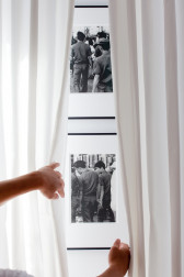 Two black and white photographs of various men casually holding hands are mounted one on top of the other on a white gallery wall behind semi-translucent white curtains. Two hands draw the curtains back to reveal the images.