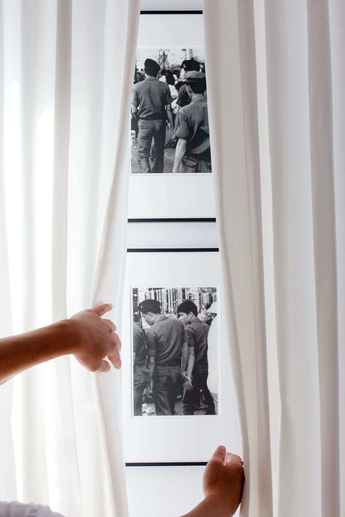 Two black and white photographs of various men casually holding hands are mounted one on top of the other on a white gallery wall behind semi-translucent white curtains. Two hands draw the curtains back to reveal the images.