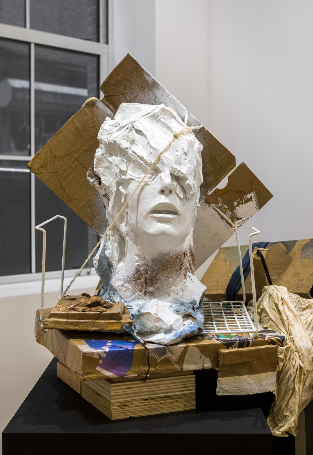 On a black platform, assorted pieces of wood, cardboard, and wire prop up a disembodied humanoid head. The figure’s head is misshapen, as if melting, obscuring the eyes but leaving the nose and open mouth visible.