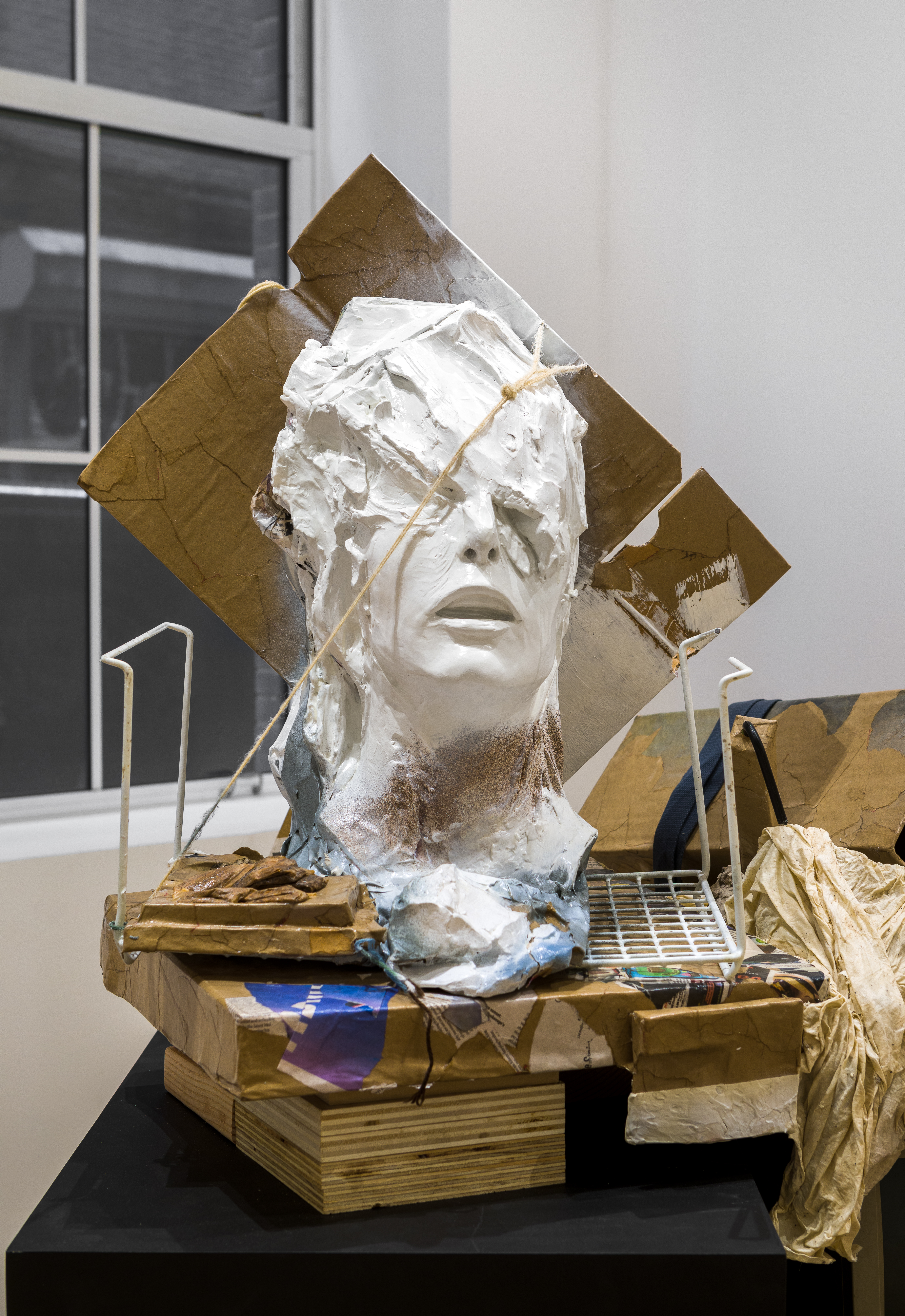 On a black platform, assorted pieces of wood, cardboard, and wire prop up a disembodied humanoid head. The figure’s head is misshapen, as if melting, obscuring the eyes but leaving the nose and open mouth visible.