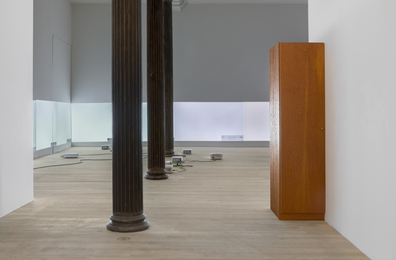 Installation view of a gallery filled with projectors arranged on the floor around three large black columns in the middle of the room. Each projector is projecting a bright white test screen onto white walls, aligned so all play at the same height. A wooden wardrobe stands in the foreground of the image against a white wall.