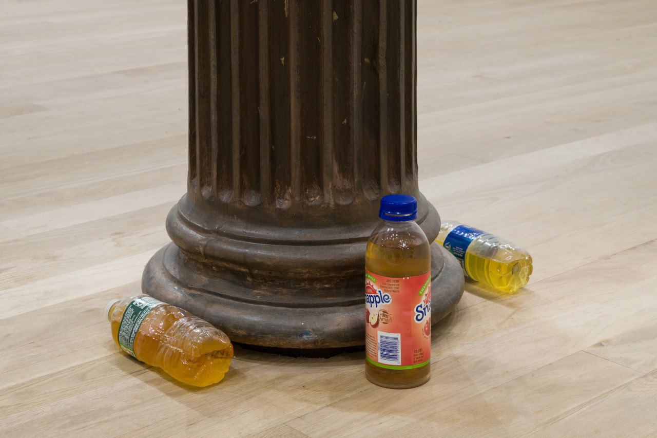 Three unmatching bottles filled with urine surround the base of a black column. Two lie horizontal, one stands upright.