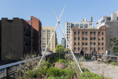 On an elevated walkway that overlooks a cityscape, three wooden rods form a teepee in a green space. A white rope hangs from where the structure is tied together, the other end suspends a large rock very close to the ground.