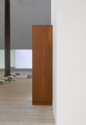 A wooden wardrobe stands against a white wall. In the background, four projectors sit on the floor amid one black column. Two white test patterns are projected on white walls.