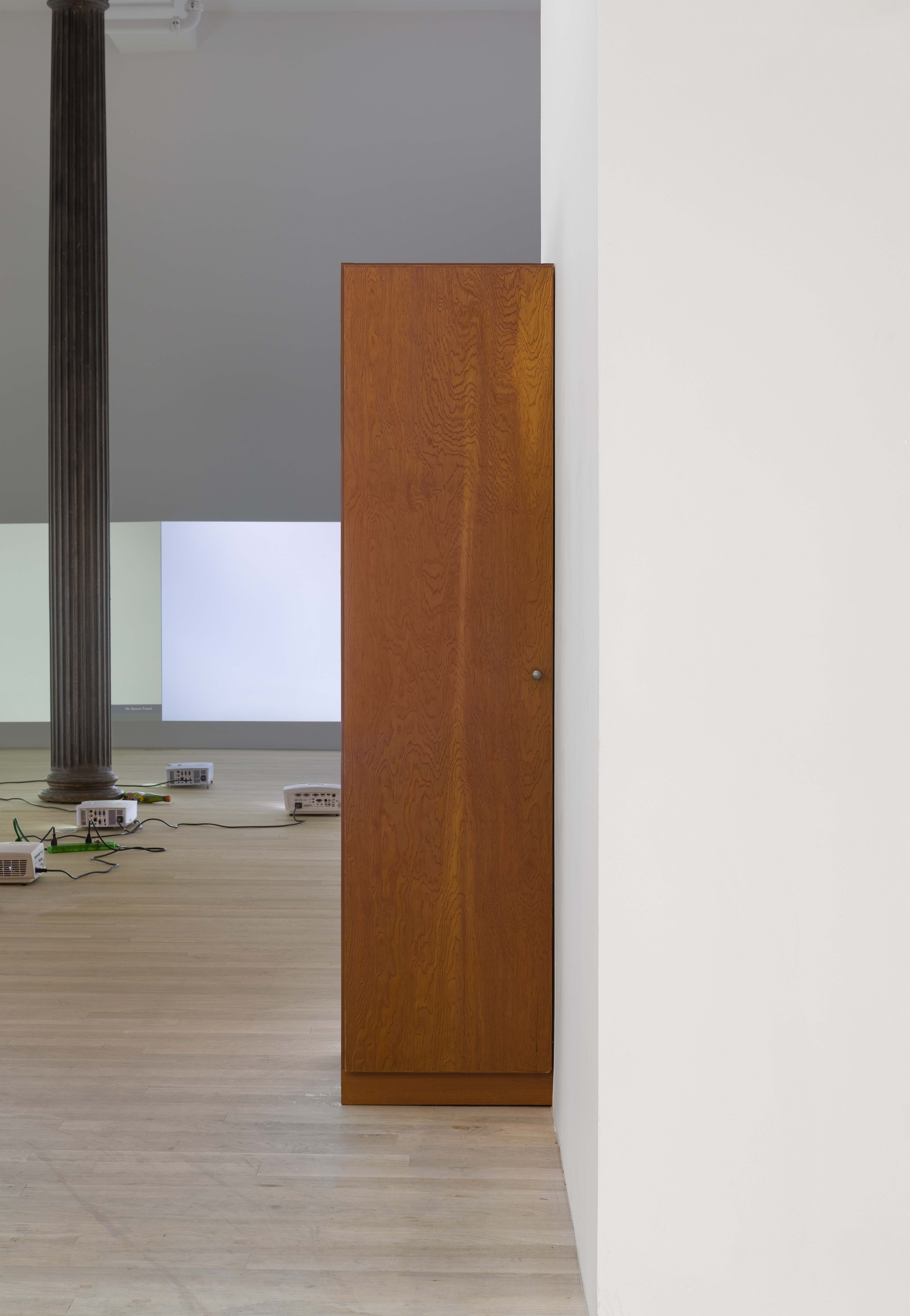 A wooden wardrobe stands against a white wall. In the background, four projectors sit on the floor amid one black column. Two white test patterns are projected on white walls.