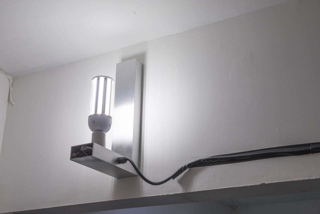 In the corner of a room, close to the ceiling, a white L-shaped wall mount holds an LED lightbulb upright. A black wire connected to the lamp is taped to the wall with black tape and disappears off to the right of the image.