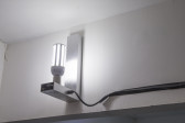 In the corner of a room, close to the ceiling, a white L-shaped wall mount holds an LED lightbulb upright. A black wire connected to the lamp is taped to the wall with black tape and disappears off to the right of the image.