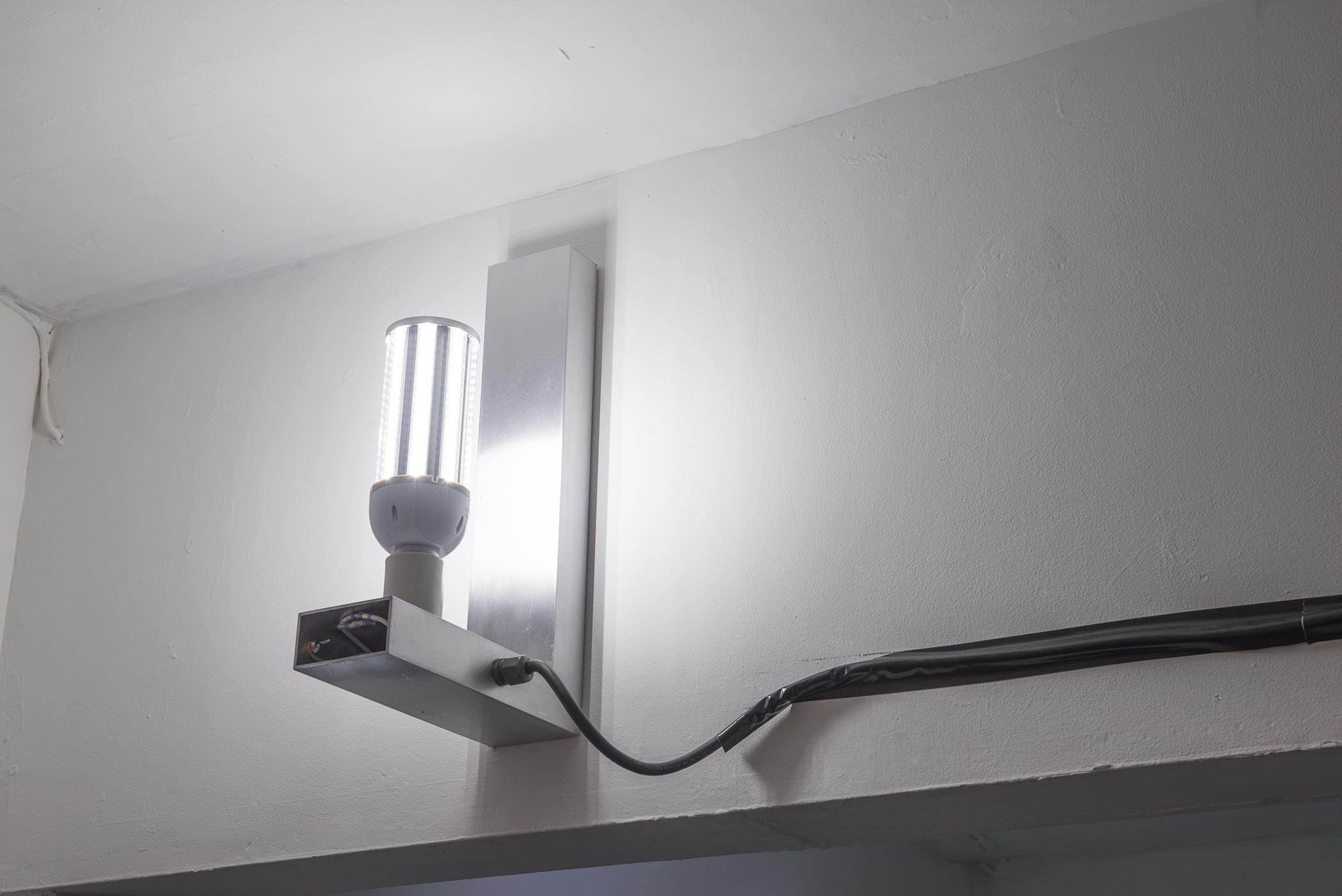 In the corner of a room, close to the ceiling, a white L-shaped wall mount holds an LED lightbulb upright. A black wire connected to the lamp is taped to the wall with black tape and disappears off to the right of the image.