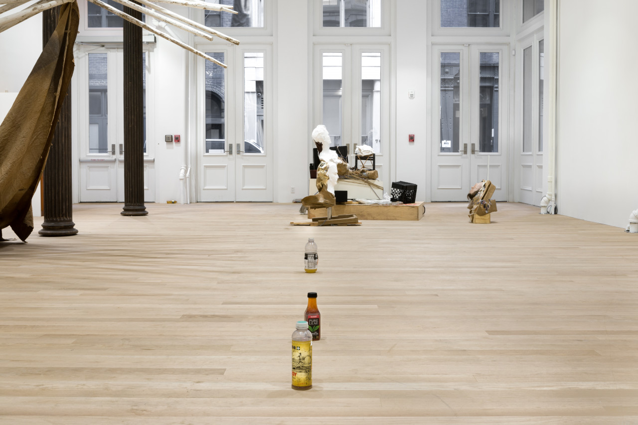 Three unmatching plastic bottles filled with urine are sitting in a line on a wood floor. In the background, a mixed-media installation piece sits in front of the gallery doors. Wooden poles project into the frame through two standing columns.
