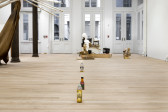 Three unmatching plastic bottles filled with urine are sitting in a line on a wood floor. In the background, a mixed-media installation piece sits in front of the gallery doors. Wooden poles project into the frame through two standing columns.