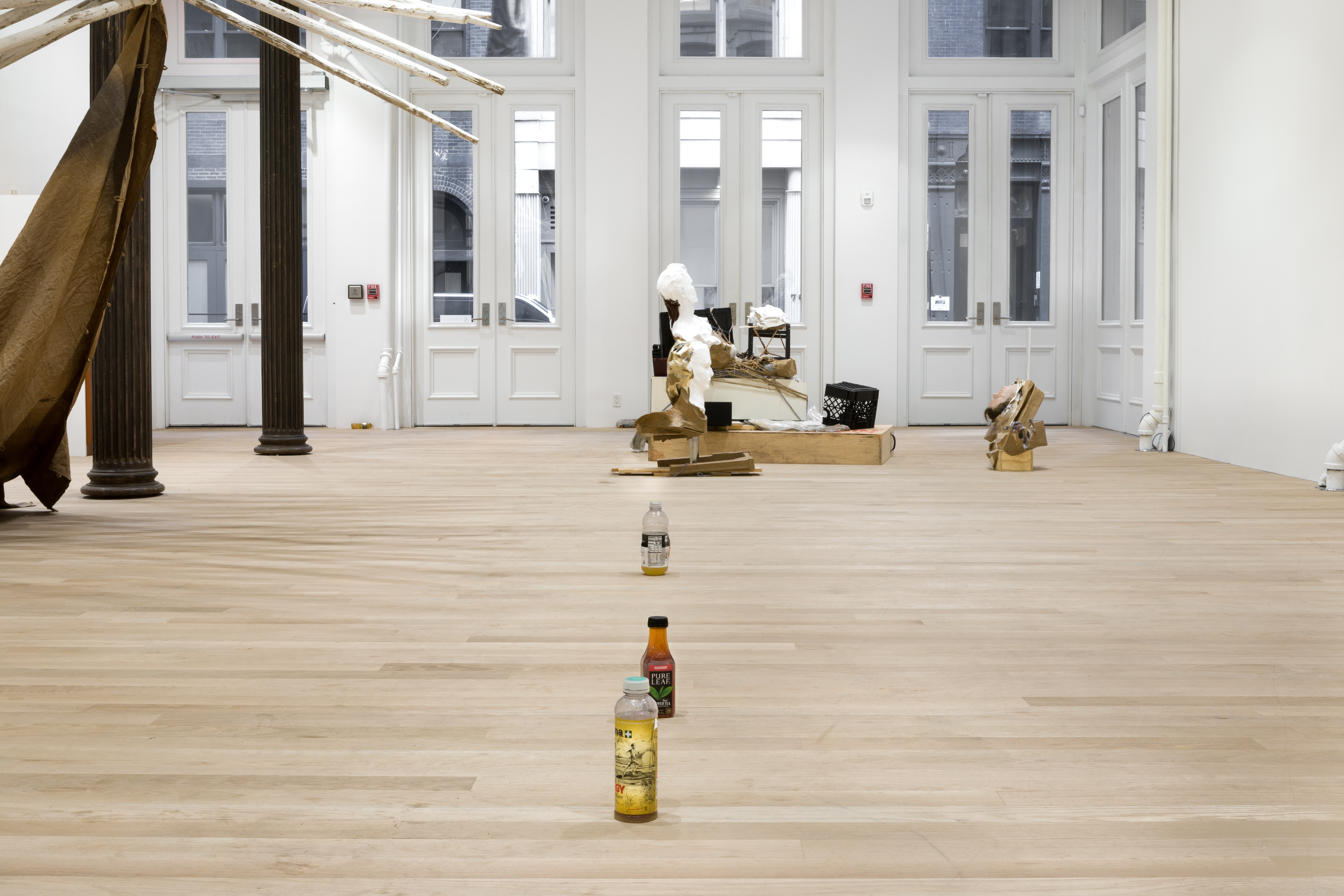 Three unmatching plastic bottles filled with urine are sitting in a line on a wood floor. In the background, a mixed-media installation piece sits in front of the gallery doors. Wooden poles project into the frame through two standing columns.
