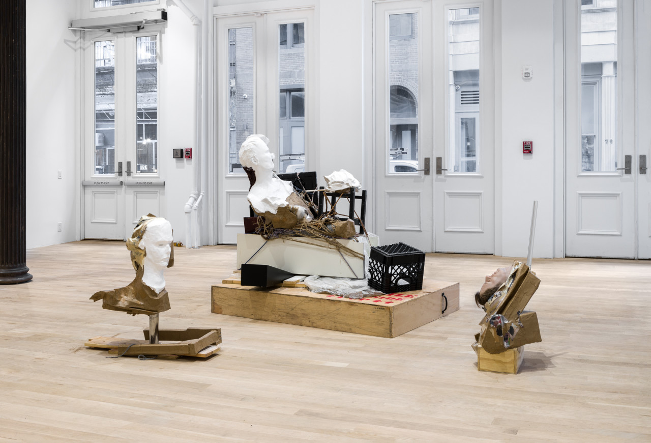 Three sculptures grouped together in the corner of a gallery, with a street in Manhattan visible through large windows at the rear of the room. The largest sculpture stands behind the other two; clustered pieces of wood, string, cardboard, paper, metal, and plastic are propped up, arranged on top of one another, all standing on a large cardboard base. A humanoid white bust lies on top of the structure, its face cut in half. The two smaller sculptures also depict humanoid faces - one in white, the other with skin and hair - and both appear to grow out of masses of misshapen cardboard and wood standing on the floor.