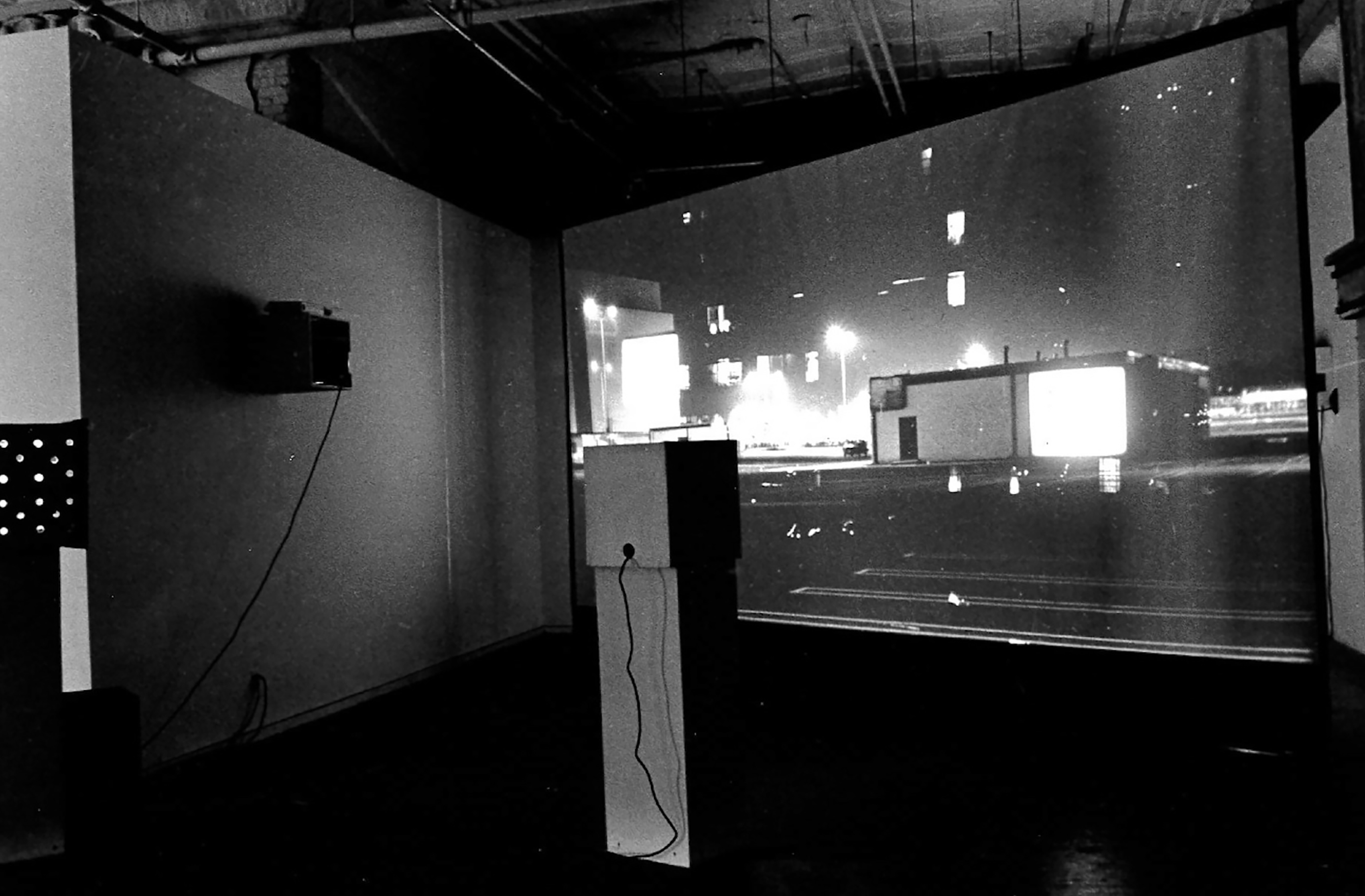 A video projection on a scrim of an urban environment with a store and a park lot visible with buildings in the background. A standing box with a wire coming from the back is visible and another box with a wire from the back is visible on the right wall. All of this is set up in a gallery setting.